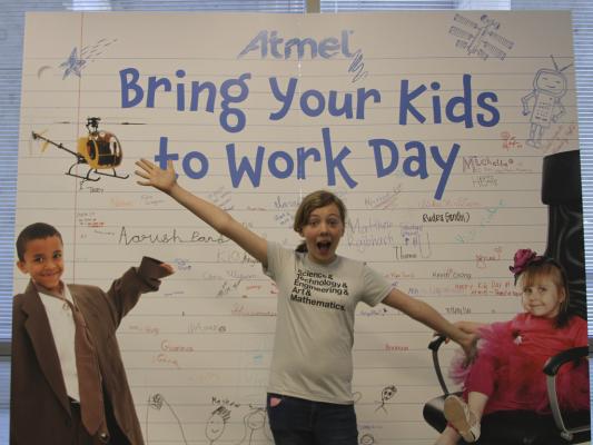 Sylvia poses in front of a giant poster for Atmel bring your kids to work day.