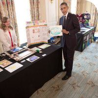 Sylvia standing behind her science fair table and her WaterColorBot while President Barack Obama displays the White House logo drawn by the bot.